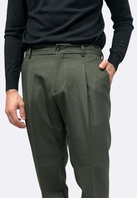 Pantaloni verde scuro con due pieghe, passanti per cintura e tasche laterali. Il materiale sembra liscio con una vestibilità sartoriale. Abbinati a una maglietta nera.