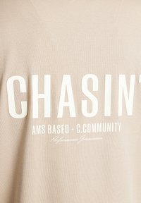 Beige sweatshirt med stor hvid tekst, der læser "CHASIN'" og "AMS BASED - C.COMMUNITY." Glat tekstur, afslappet design og minimal detaljer.