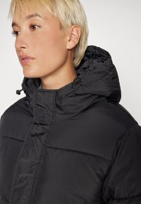 Veste en duvet noire avec un col haut et une capuche attachée. Présente une texture matelassée et des cordons de serrage ajustables à la capuche.