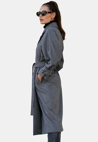 Szary płaszcz z miękkiego materiału, z talią przewiązaną paskiem, długimi rękawami i bocznymi kieszeniami. Uzupełniony czarnymi okularami przeciwsłonecznymi i wysokimi czarnymi kozakami.