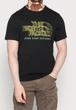 Mężczyzna ubrany w czarną koszulkę The North Face z żółtym logo w formie siatki i napisem "Never stop exploring" oraz brązowe spodnie typu cargo z kieszeniami.