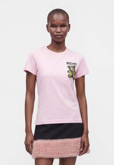 Γυναίκα που φοράει ένα ανοιχτό ροζ t-shirt Moschino με γραφικό αρκουδάκι και μαύρη μίνι φούστα με ροζ χνουδωτό τελείωμα στο κάτω μέρος.