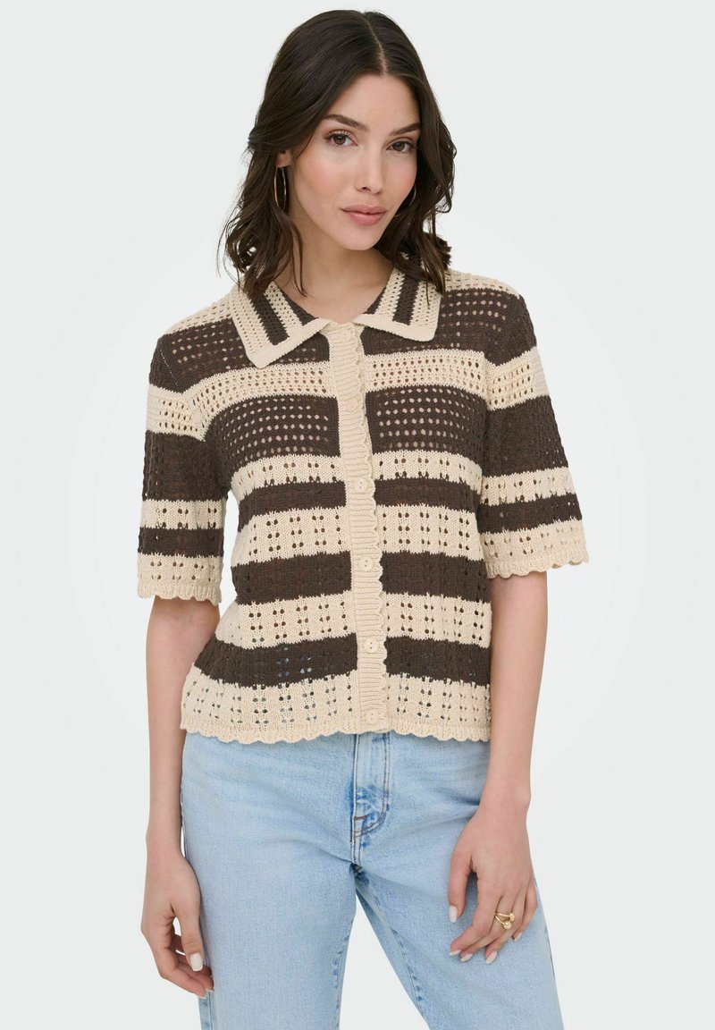Femme aux cheveux mi-longs et foncés portant un gilet court en crochet rayé crème et marron à manches courtes, ainsi qu'un jean bleu clair, debout devant un fond blanc.