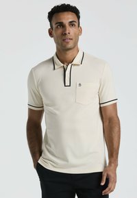 Polo beige avec des bordures noires et trois boutons, doté d'une poche poitrine avec un logo. Texture lisse et design ajusté.