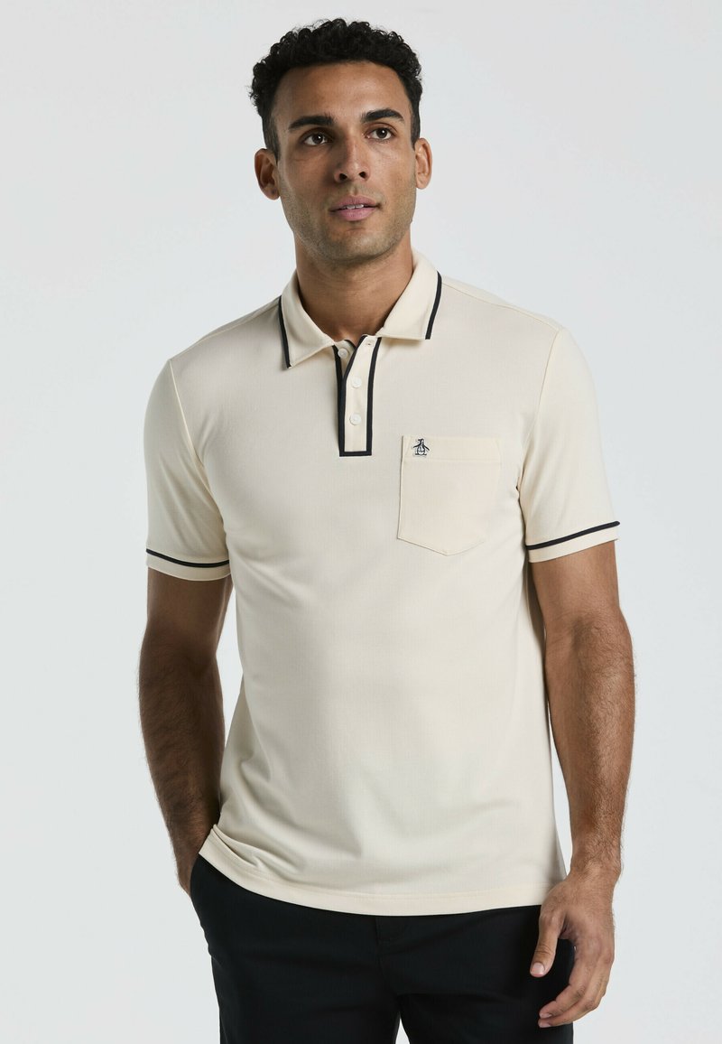 Polo beige avec des bordures noires et trois boutons, doté d'une poche poitrine avec un logo. Texture lisse et design ajusté.