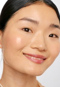 Close-up van een persoon met een gladde, lichte huid; donker, steil haar; natuurlijke makeup; geaccentueerde wangen; en een roze glans op de lippen. Neutrale achtergrond.
