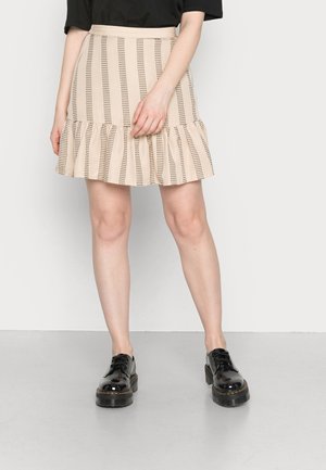 Saias bege com folhos, riscas verticais e detalhes recortados, combinadas com sapatos pretos de couro verniz com atacadores e uma blusa preta solta.