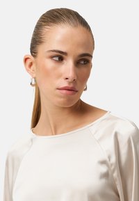 Světle béžový saténový top s kulatým výstřihem, hladkou texturou a dlouhými rukávy. Modelka má stříbrné kruhové náušnice.