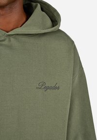 Oliwkowa bluza z kapturem o miękkiej fakturze. Posiada przednią kieszeń typu kangur oraz haftowane ciemnoniebieskie słowo "Pegador" na klatce piersiowej.