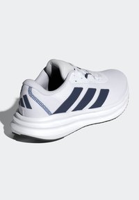 Chaussure de sport blanche avec des bandes noires, semelle coussinée et texture en mesh, vue d’un angle latéral arrière sur un fond uni.