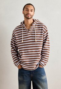 Brun og lyseblå stripet hettegenser med frontlomme, snor og en avslappet passform, sammen med blå jeans. Strukturert stoff.