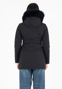 Giubbotto puffer nero con cappuccio e rifinitura in pelliccia sintetica. Presenta una vita aderente e maniche lunghe. Indossato sopra jeans di denim blu larghi.