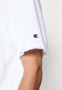 Άσπρο βαμβακερό T-shirt με ακατέργαστο τελείωμα στα μανίκια και μικρό κεντημένο λογότυπο σε μαύρο, κόκκινο και άσπρο στο μανσέτα.