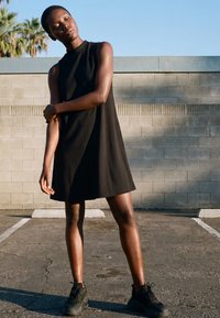 Fiatal nő fekete ujjatlan ruhában és fekete tornacipőben áll a napsütötte járdán, zárt szemmel megérinti a karját, téglafal közelében.