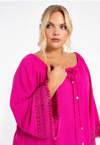 Tunique fuchsia avec un col carré, un lien à l'avant et des boutons. Présente des accents en dentelle crochetée au niveau des manches pour plus de texture et de détail.