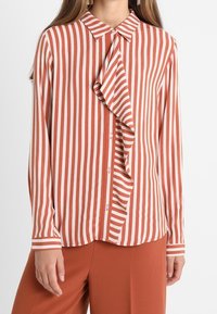 Randig blus i rostfärgad och vit, med en volang framknäppning, krage och knappstängning. Långa ärmar med randiga muddar.