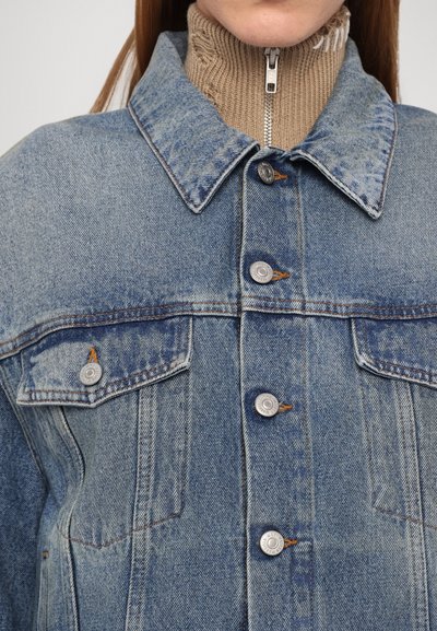 Denim jacket in light wash with pointed collar, silver button closures, and two chest pockets. Features tan ribbed turtleneck underneath.