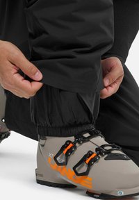 Schwarze wasserdichte Hose mit elastischen Bündchen, die an den Knöcheln verstellbar ist, getragen über grauen Skischuhen mit schwarzen und orangenen Schnallen sowie Branding.