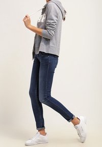 Sudadera gris con cremallera y letras blancas, combinada con jeans ajustados azul oscuro y zapatillas blancas. Tiene capucha y cordones.