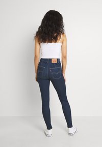 Jeans ajustados de tiro alto en azul oscuro con un parche de cuero, bolsillos traseros y costuras contrastantes, combinados con una camiseta sin mangas blanca y zapatos blancos.