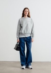 Femme debout contre un mur blanc, portant un sweat-shirt gris, un jean bleu à jambes larges, des baskets blanches et tenant un petit sac à main noir.