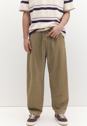 Persoană care poartă pantaloni largi kaki cu talie reglabilă prin șnur, un tricou în dungi și adidași maro închis cu tălpi și șireturi albe.