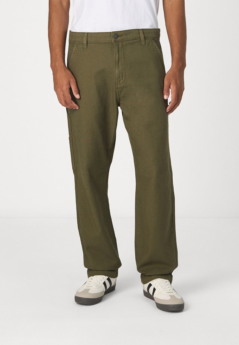 Pantalones verde oliva hechos de una tela resistente, con un diseño de pierna recta, cierre de botón y bolsillos laterales. Llevados con zapatos blancos y grises.