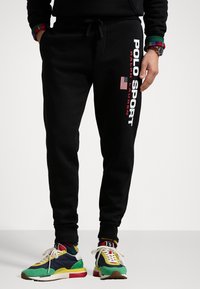 Černé tepláky s nápisy "POLO SPORT" a "RALPH LAUREN" v bílé a červené barvě, s akcentem americké vlajky na levé noze.