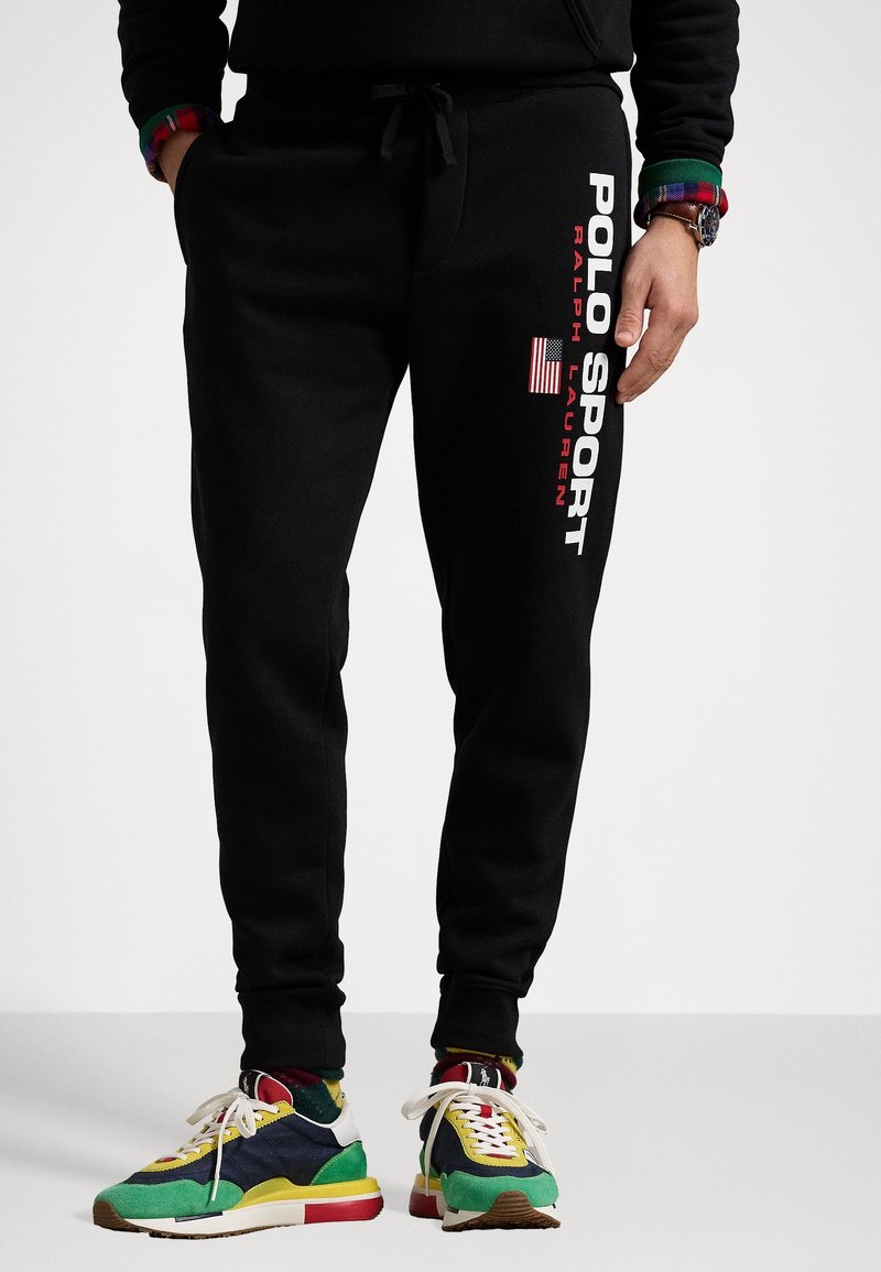 Černé tepláky s nápisy "POLO SPORT" a "RALPH LAUREN" v bílé a červené barvě, s akcentem americké vlajky na levé noze.