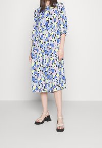 Florale midi-jurk met blauwe, paarse en groene patronen, met 3/4 mouwen en een gerimpelde taille. Gepaald met beige bandjeshakken.