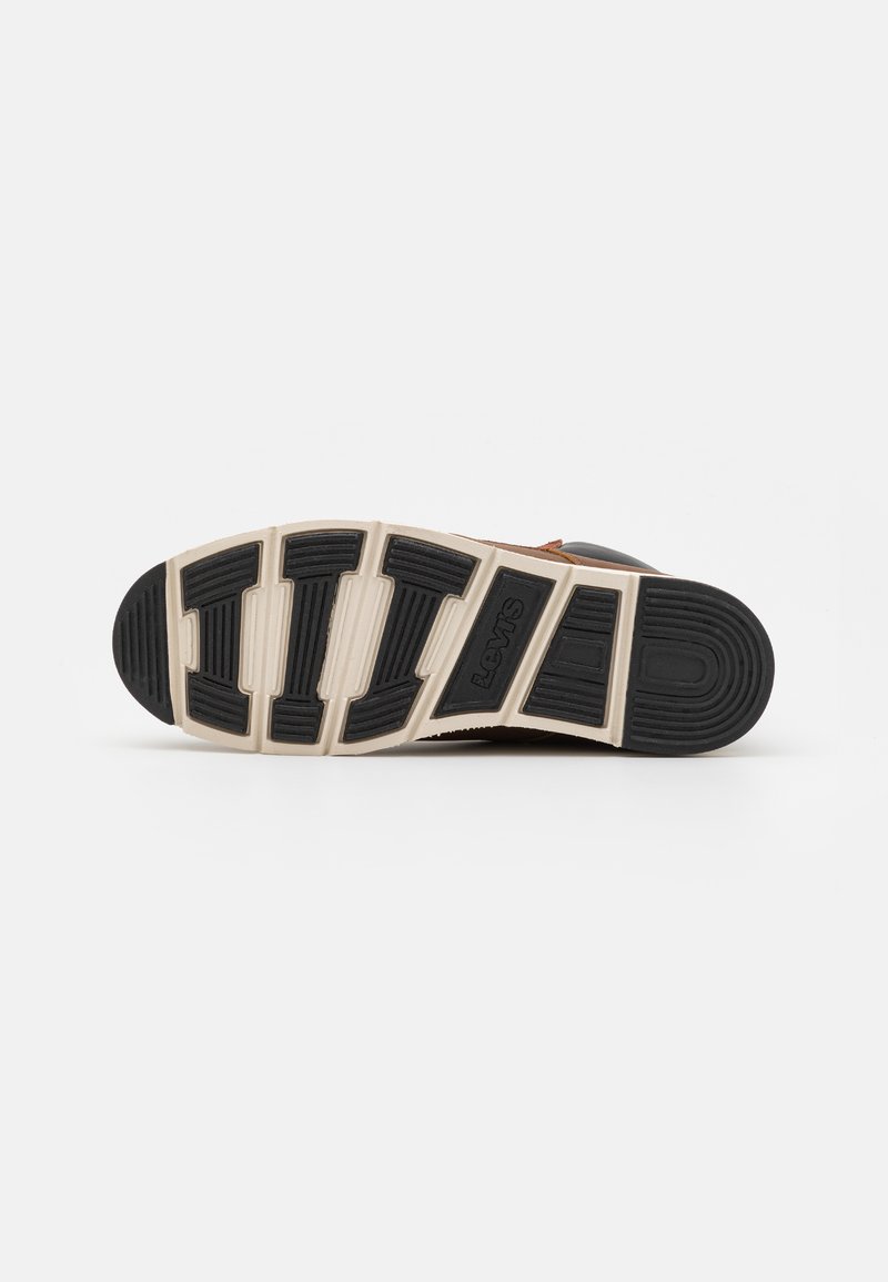Suela de goma negra con patrón de textura, entresuela crema y acentos marrones. Vista inferior de una zapatilla casual, con un diseño resistente.