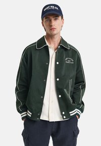 Giacca da baseball verde con piping bianco, bottoni e logo. Indossata sopra una camicia chiara con bottoni. Presenta anche un cappellino scuro. Design casual.