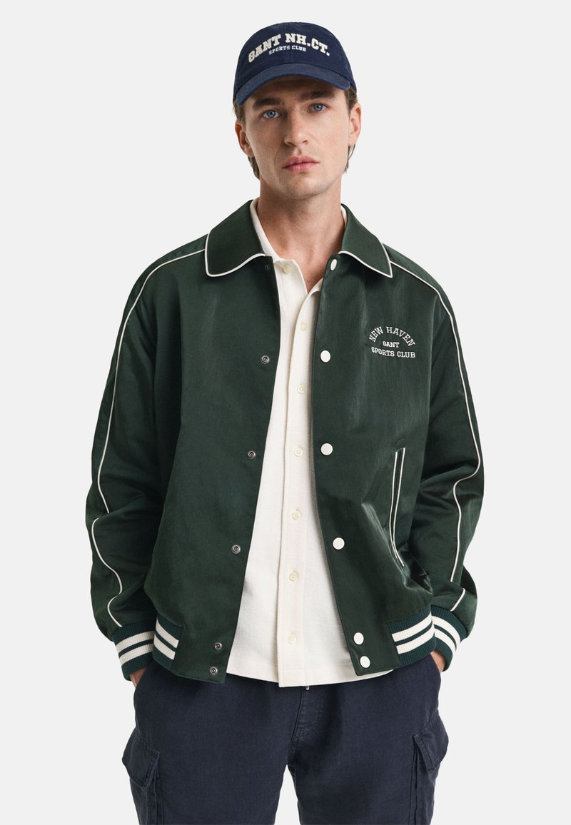 Grüne Baseballjacke mit weißen Abnähern, Knöpfen und Logo. Über einem hellen Hemd mit Knopfleiste getragen. Zusätzlich verfügt sie über eine dunkle Kappe. Lässiges Design.