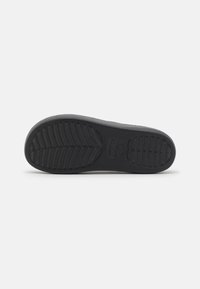 Semelle de chaussure en caoutchouc noir avec un motif de grip texturé, présentant des stries définies et une marque estampillée au centre. Forme plate et large.