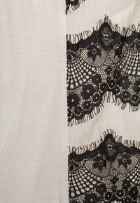 Witte stof met drie horizontale banden van intrigerende zwarte kant met bloemmotieven, afgewerkt met rafelige details.