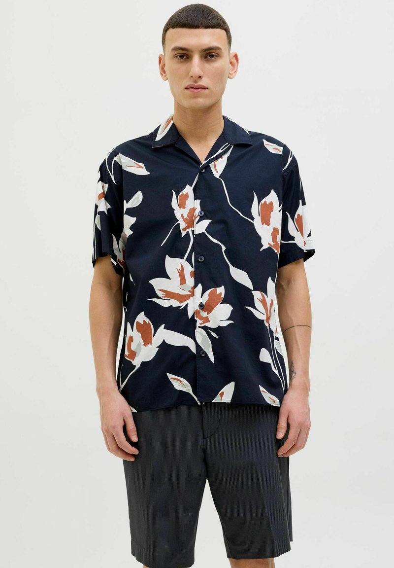 Camisa de manga corta en azul marino con un gran patrón floral blanco y naranja; presenta un cierre de botones en la parte delantera y un cuello, hecha de tejido ligero.