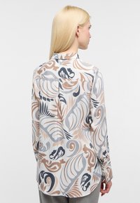 Donna bionda vista da dietro, che indossa una camicia a maniche lunghe con bottoni, dai motivi astratti a foglia beige, grigio e nero.