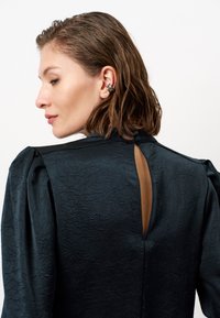 Schwarze Textbluse mit gepufften Ärmeln und einem Schlüsselaugenverschluss auf der Rückseite. Sie besteht aus einem glatten, leicht zerknitterten Stoff und hat einen Knopfverschluss am Hals.