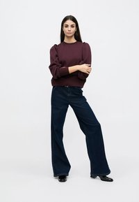 Burgunderfarget genser med pufferm, kombinert med vide mørke denimbukser. Glatt tekstur, minimalistisk design. Langt mørkt hår, stående stilling.