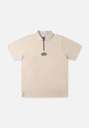 Beige poloshirt med korte ærmer, lilla lynlås og broderet logo på brystet. Har en standard krave og et minimalistisk design.