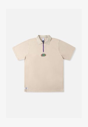Beige poloshirt med korte ærmer, lilla lynlås og broderet logo på brystet. Har en standard krave og et minimalistisk design.