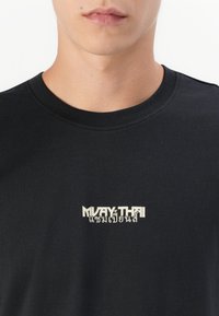 Close-up of a person wearing a black t-shirt with a white "MUAY THAI" and Thai script logo centered on the chest.