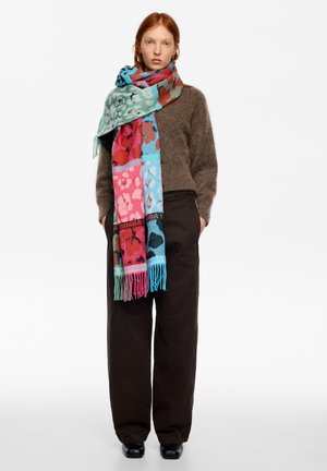 Bufanda multicolor en azul, rosa y verde con un patrón floral y bordes con flecos, llevada sobre un suéter marrón y pantalones oscuros sueltos.