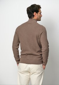 Brauner Strickpullover mit hohem Kragen, enganliegende Ärmel und strukturiertem Muster, kombiniert mit cremefarbenen Hosen für den Kontrast.
