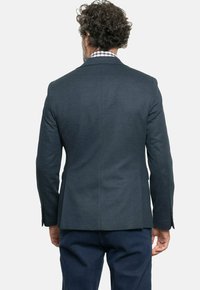 Blazer azul oscuro hecho de tejido texturizado, con una sola abertura en la parte trasera y solapas con muescas, usado sobre una camisa de cuadros.