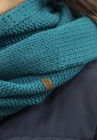 Sciarpa lavorata a maglia color teal con un motivo testurizzato e un'etichetta in pelle. La sciarpa è sovrapposta a una giacca scura.