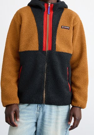 Hombre con chaqueta Columbia de forro polar marrón y negro con cremallera roja y detalles en los bolsillos, combinada con vaqueros azul claro.