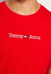 Červené tričko s bílým nápisem "Tommy Jeans" a malým obdélníkovým logem v námořnické, bílé a červené barvě umístěným na středu hrudi.