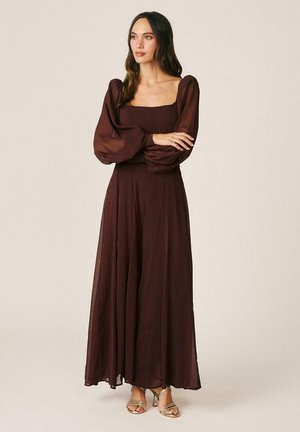 Mujer con un vestido largo marrón oscuro con mangas largas abullonadas, de pie con los brazos cruzados, calzando sandalias de tacón de color claro.