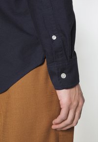 Camicia button-up blu navy con tessuto testurizzato, caratterizzata da bottoni argento ai polsini e orlo curvo, abbinata a un indumento intrecciato color beige.